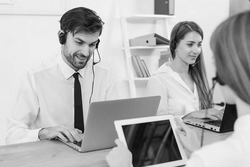 Two customer service representatives wearing headsets sit at a desk, working on laptops for emergency IT help desk support. Another person in the foreground holds a tablet in the bright, modern office environment.