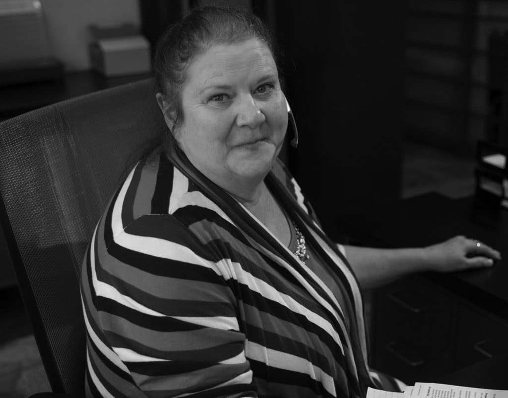pavelcomm-hipaa-new-1024x803-11 A woman with light skin and brown hair, wearing a blue, teal, and white striped top, sits at an office desk with papers and a headset, smiling at the camera—demonstrating a professional setting focused on HIPAA compliance.