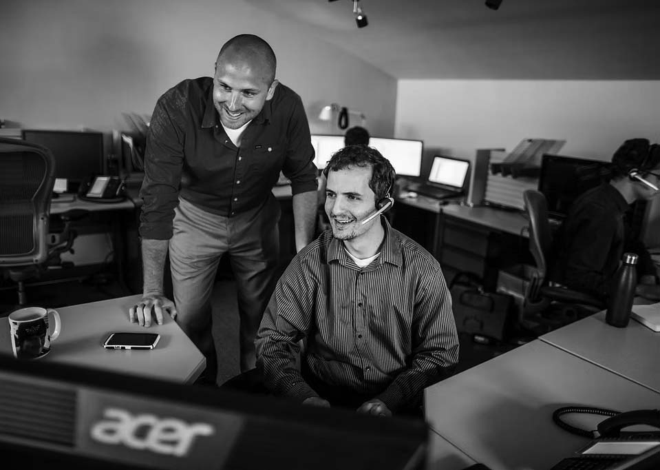 support-services Two men in an office, one standing and one sitting at a desk, both smiling and looking at a computer monitor. The seated man wears a headset, reflecting the Support provided by Pavelcomm. Other workers with headsets are visible in the background.