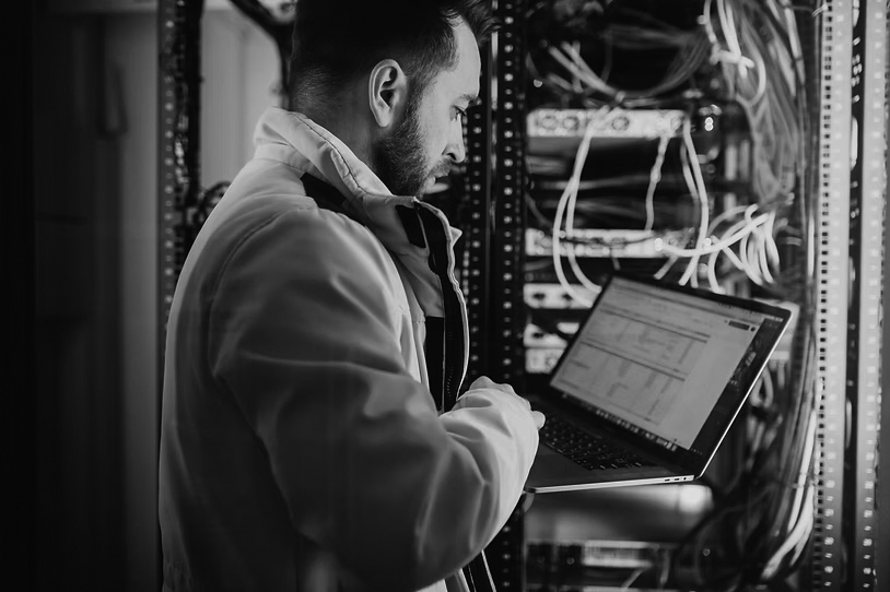 A man in a yellow jacket works on a laptop while standing next to a server rack filled with cables and equipment in a data center.