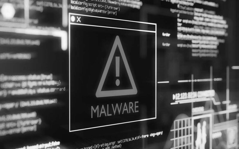 A digital screen displays a warning triangle with an exclamation mark and the word MALWARE in bold, surrounded by glowing lines and computer code, representing a cybersecurity threat and risk of data breaches.