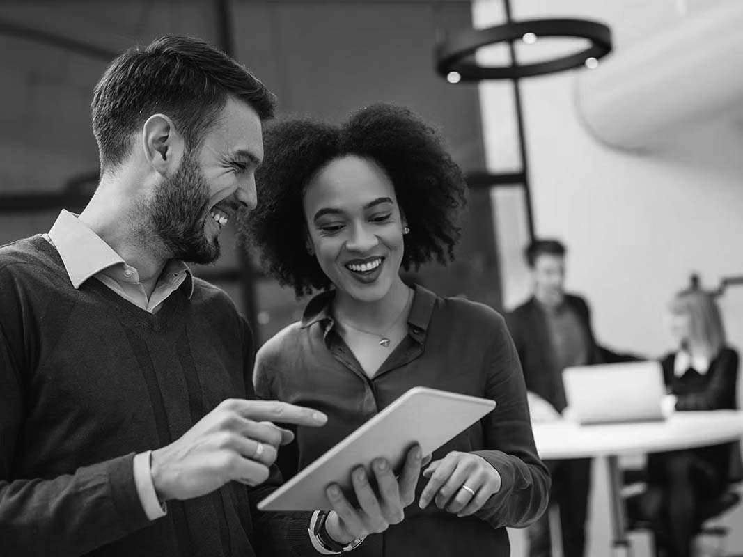 Two coworkers, a man and a woman, smiling and looking at a tablet together in a modern office—perhaps exploring the benefits of cloud communications. In the background, two other colleagues work at a desk with laptops.