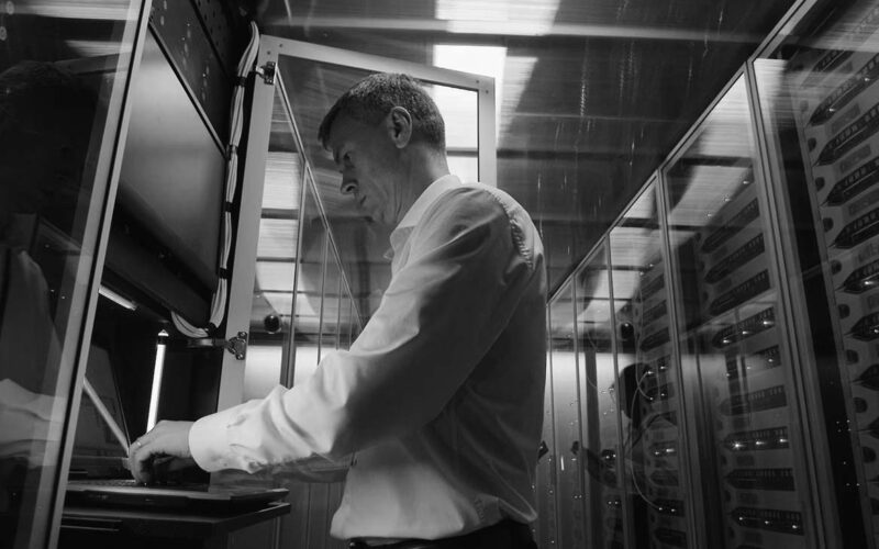 A man in a white shirt stands in a server room, working on a laptop mounted in a server rack. Surrounded by blinking lights, he focuses on cyber security to help the data center regain control after a potential cyberattack.