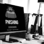 A computer screen displays an IT security phishing warning with a cartoon robot and the words PHISHING plus a red SCAN NOW button. The desk has office supplies, a coffee cup, and a red toy with teeth to help prevent cyber attacks.