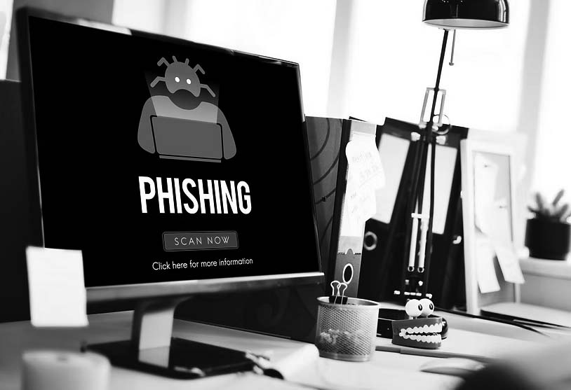 A computer screen displays an IT security phishing warning with a cartoon robot and the words PHISHING plus a red SCAN NOW button. The desk has office supplies, a coffee cup, and a red toy with teeth to help prevent cyber attacks.