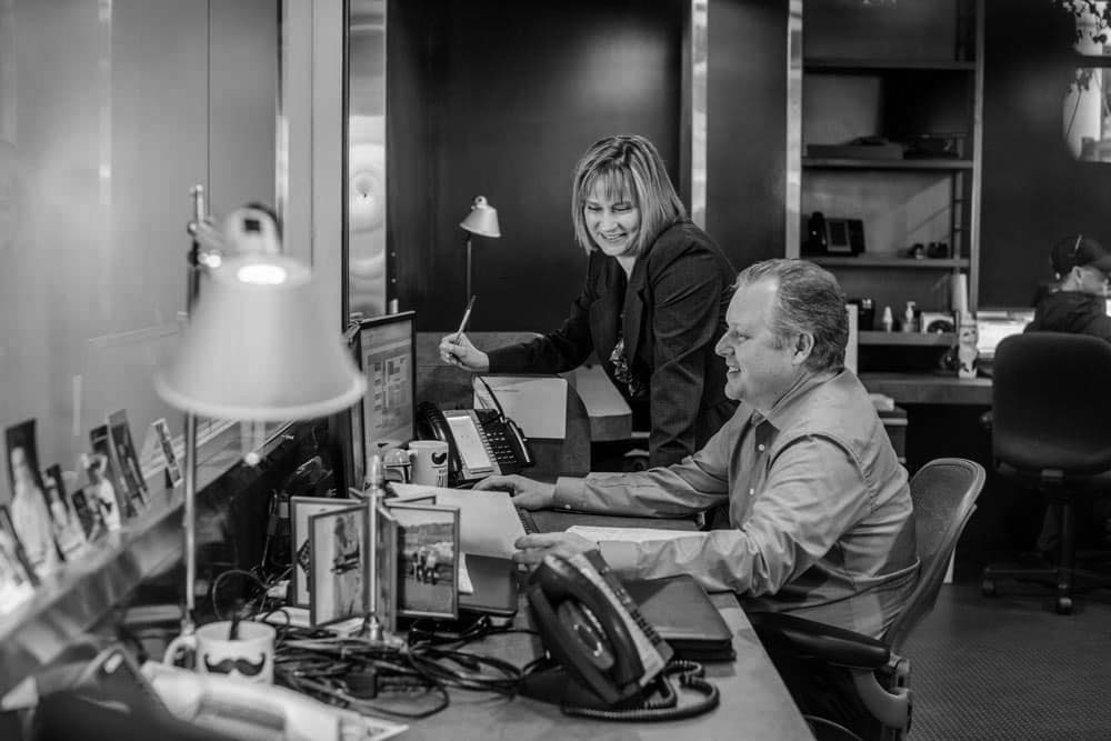 H139363 Two people in business attire work at a desk with computers in a modern office, collaborating on customer solutions. One types while the other stands, smiling and holding a pen. Office equipment and framed photos are visible on the desk.