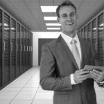 A smiling man in suit standing in a server room, looking into the camera while holding a tablet.