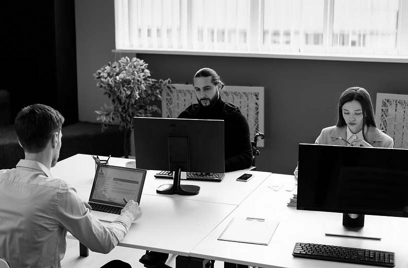 An office setting with several people sitting around laptop and desktop computers thinking and looking at their workstations.