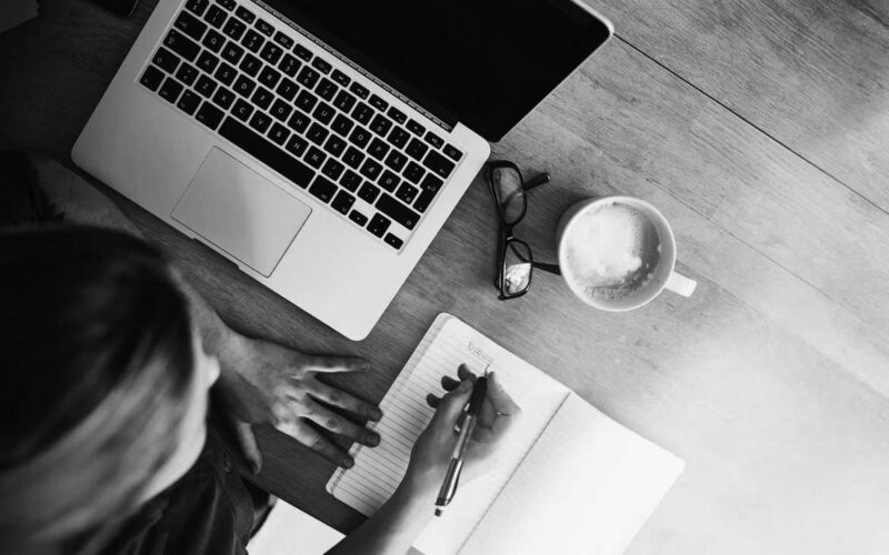 A person writes in a notebook at a wooden desk with a laptop, eyeglasses, and a cup of coffee nearby—perhaps planning steps to become PCI compliant. The scene is lit by natural light from the side.