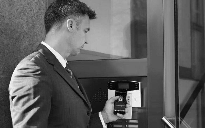 A business professional in a suit holds a smartphone displaying a home security app up to a smart access control panel at a building entrance, showcasing seamless IoT integration while attempting to unlock the door.