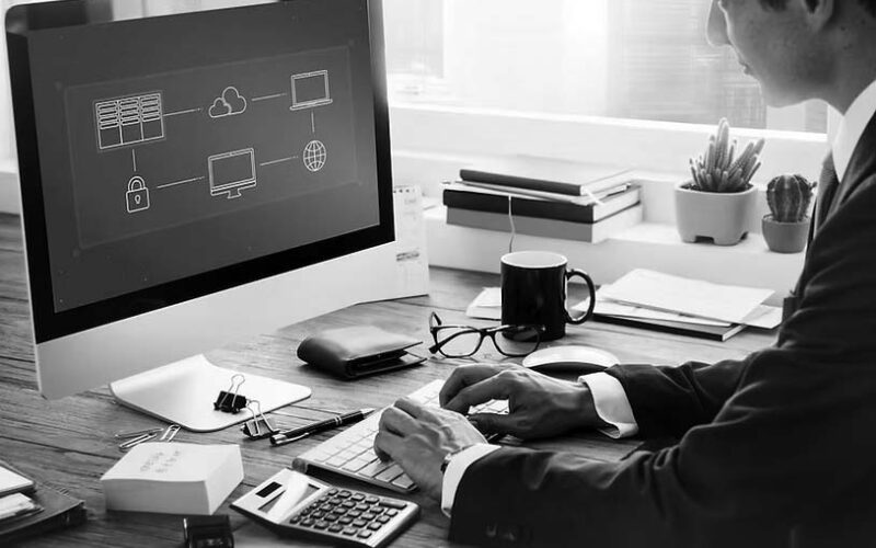 A person in a suit sits at a desk using a computer displaying a network diagram, researching Migration & Deployment Solutions from Pavelcomm. The desk has books, a calculator, wallet, smartphone, coffee, and plant. Large windows are in the background—Portland skyline visible.