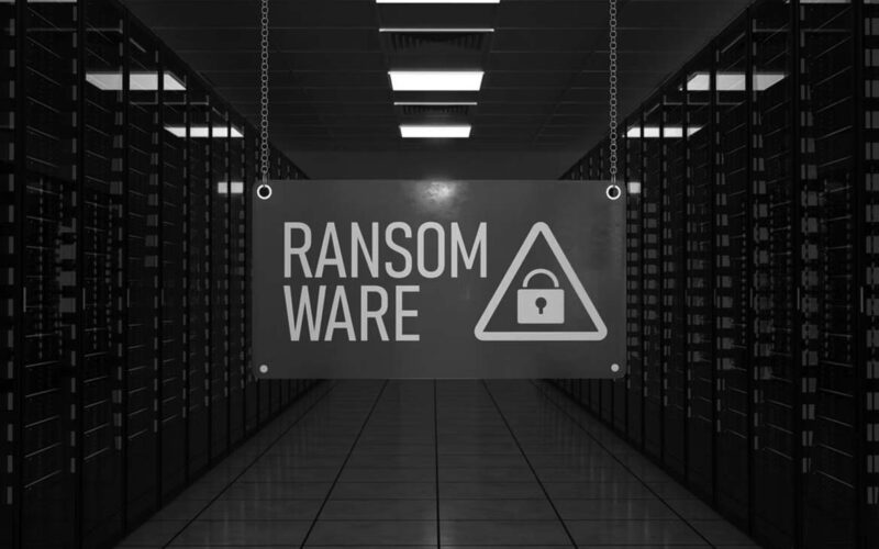 A sign reading RANSOMWARE with a lock icon hangs in a Portland server room filled with rows of servers lit by red lights, conveying the looming threat of ransomware attacks.