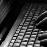 Close-up of hands typing on a laptop keyboard with lines of code visible on the screen, highlighting efforts to safeguard against ransomware and protect business security through coding activity.