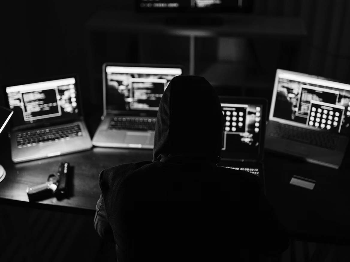 A person wearing a hood sits at a desk in Portland, surrounded by four laptops displaying code—suggesting hacking activity tied to the Dark Web. A handgun and smartphone rest on the table in the dimly lit room.