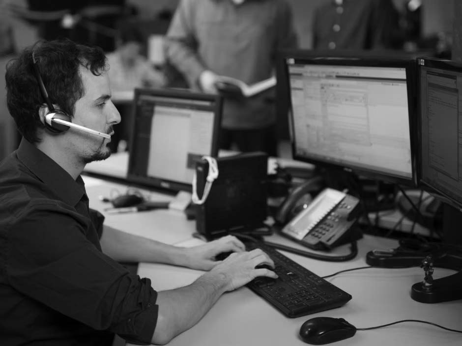 A man wearing a headset sits at a desk with three computer monitors, typing on a keyboard in an office. Other people are visible in the background, collaborating and talking, creating an optimized work environment.