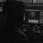 A person in a hoodie sits in front of multiple computer screens displaying code and a digital map with a target alert, holding a partially eaten sandwich in one hand. The room is dimly lit, giving a secretive atmosphere.