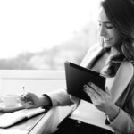 A woman sits at a table with a coffee cup, smiling while holding a tablet in one hand and writing notes for her company self-assessment in a notebook with the other. She wears a light blazer and has long hair.
