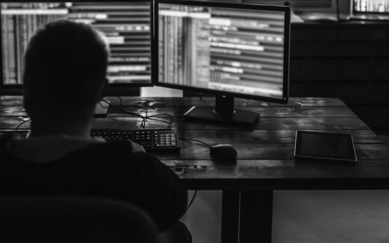 A person sits at a desk working on two computer monitors displaying lines of code related to cybersecurity in a dimly lit room. A keyboard, mouse, and tablet are also on the desk.