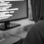 A person in a gray hoodie sits at a desk, facing a computer monitor displaying lines of code with a blue background. The scene suggests programming or cybersecurity activities—don’t wait to protect yourself against online threats.