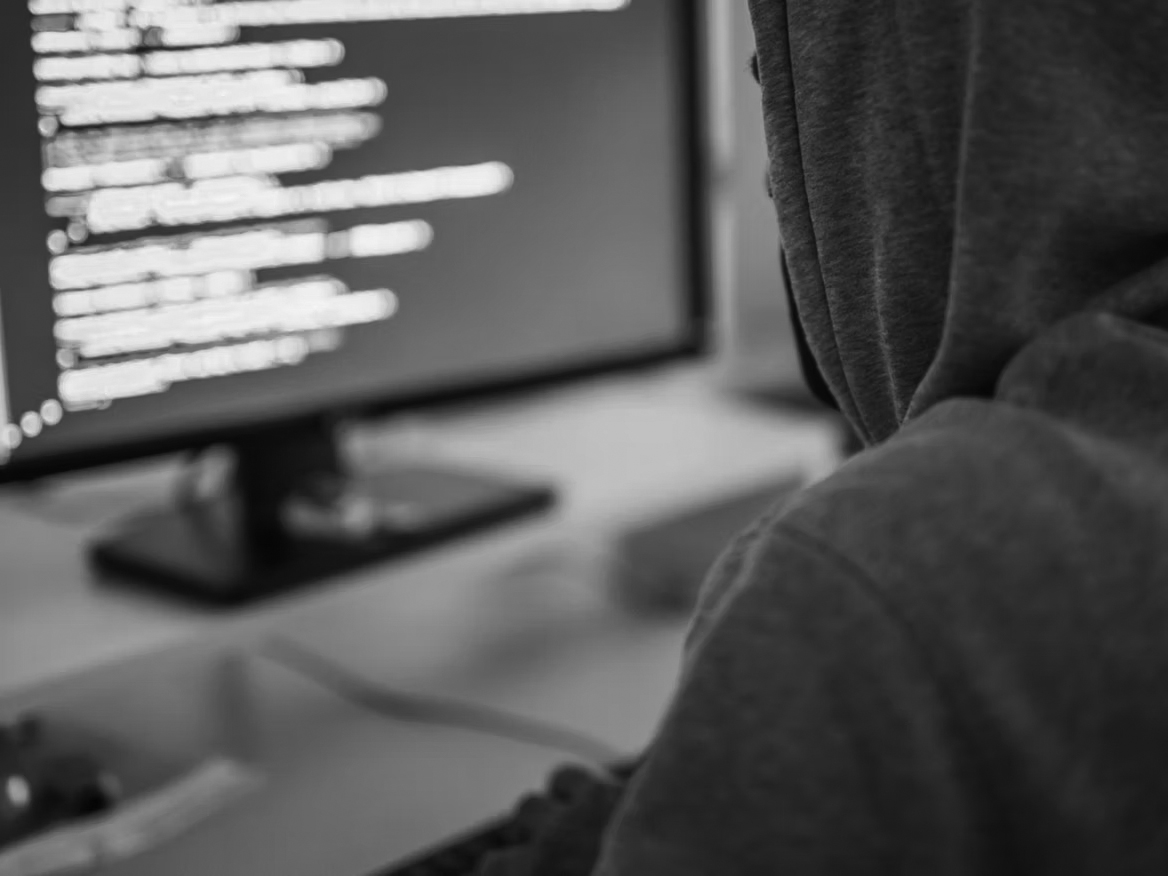 A person in a gray hoodie sits at a desk, facing a computer monitor displaying lines of code with a blue background. The scene suggests programming or cybersecurity activities—don’t wait to protect yourself against online threats.