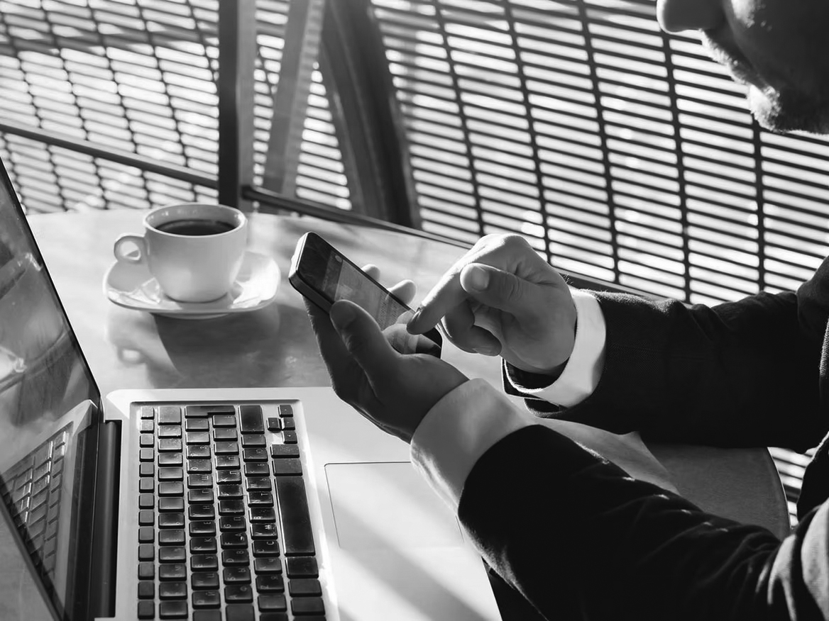 A person in a suit uses a smartphone while sitting at a table with an open laptop and a cup of coffee, sunlight streaming through window blinds—highlighting the importance of privacy and ethics in today’s connected workspaces.