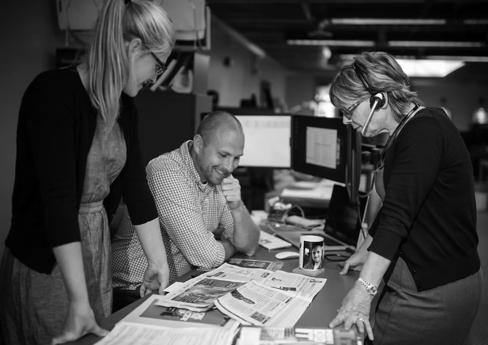 relocation-deployment-1 Three people in an office discuss printed newspapers and papers spread on a desk, exploring deployment solutions; two women stand on either side, while a man sits in the center, smiling. Computers and office supplies are visible.