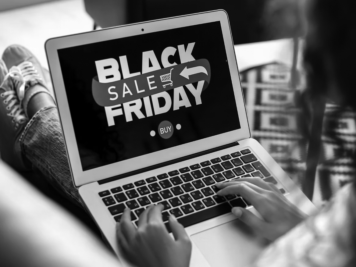 A person uses a laptop displaying a Black Friday Sale graphic with a red arrow, shopping cart icon, and Buy button, highlighting an online shopping event and the importance of safe online shopping for secure holidays.