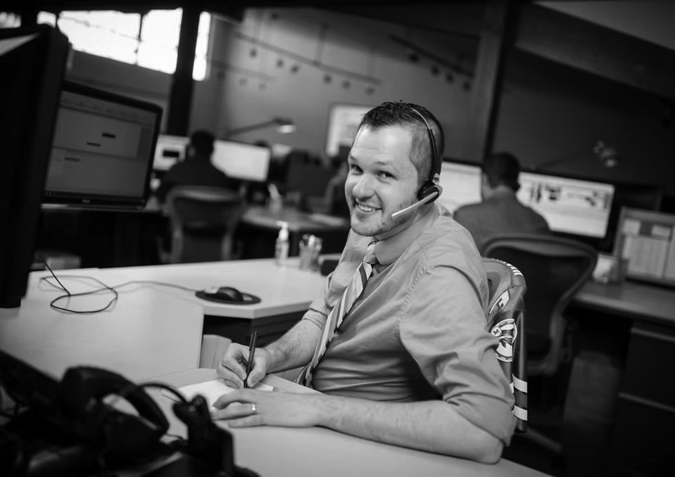server-network-solutions A smiling man wearing a headset and bright blue shirt sits at a desk in a Portland office, writing on paper while looking at the camera. Computer monitors and colleagues working on network solutions are visible in the background.