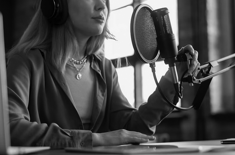 A woman wearing headphones sits at a table, speaking into a microphone on a stand. Dressed in a blue shirt and layered necklaces, she appears to be podcasting about IT Voice Solutions, gaining a competitive advantage in a studio setting.