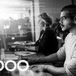 A man and a woman work at computers in an office. The man, focused in glasses and a light sweater, is surrounded by bright digital code—capturing the fast-paced environment of a Managed Services Provider working to minimize IT downtime.