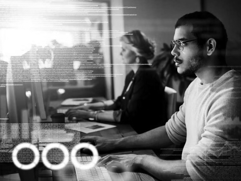A man and a woman work at computers in an office. The man, focused in glasses and a light sweater, is surrounded by bright digital code—capturing the fast-paced environment of a Managed Services Provider working to minimize IT downtime.
