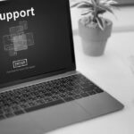 A laptop displays a screen with the word Support and a blue graphic, highlighting Microsoft 365 professional support, sitting on a white desk next to a potted plant and a stack of colored folders.