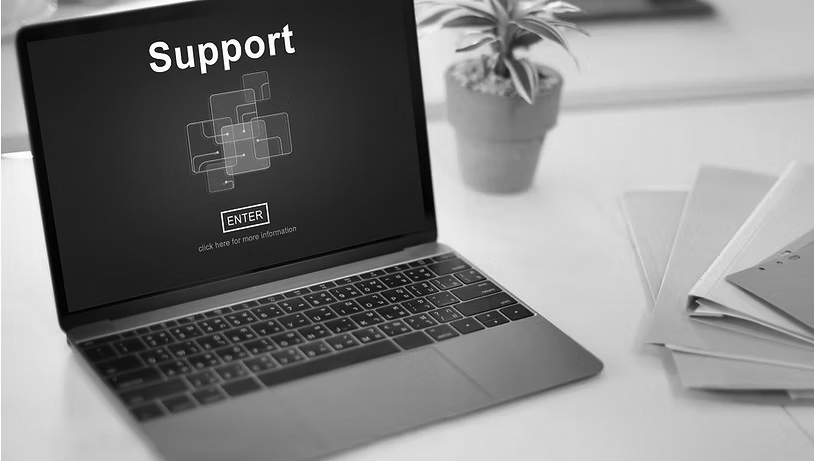 A laptop displays a screen with the word Support and a blue graphic, highlighting Microsoft 365 professional support, sitting on a white desk next to a potted plant and a stack of colored folders.