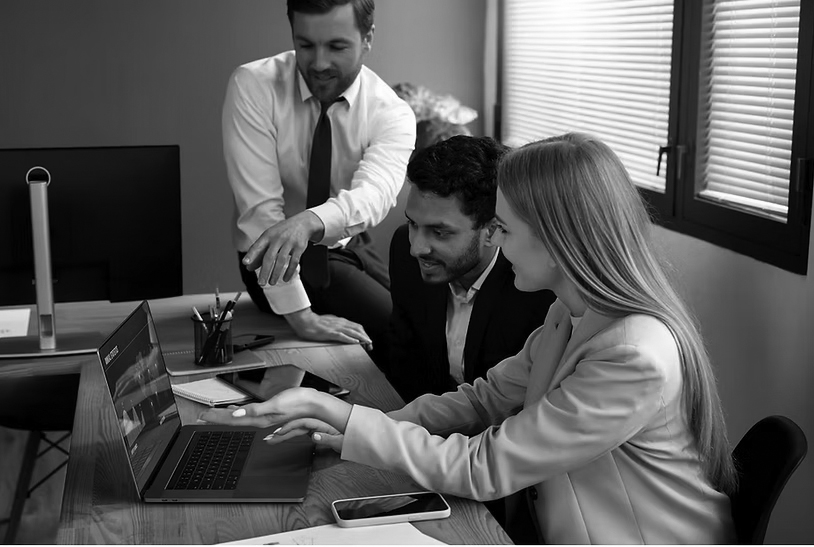 Three colleagues in business attire gather around a wooden desk, discussing IT solutions while reviewing information on a laptop. Office supplies and smartphones are visible on the desk in this modern setting, emphasizing their focus on business potential.