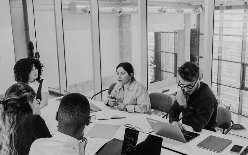 Five people sit around a conference table in a modern office, engaged in discussion about IT solutions to support non-profits. Laptops, notebooks, and papers are on the table, with large windows and glass walls visible in the background.