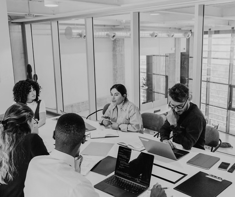 Five people sit around a conference table in a modern office, engaged in discussion about IT solutions to support non-profits. Laptops, notebooks, and papers are on the table, with large windows and glass walls visible in the background.
